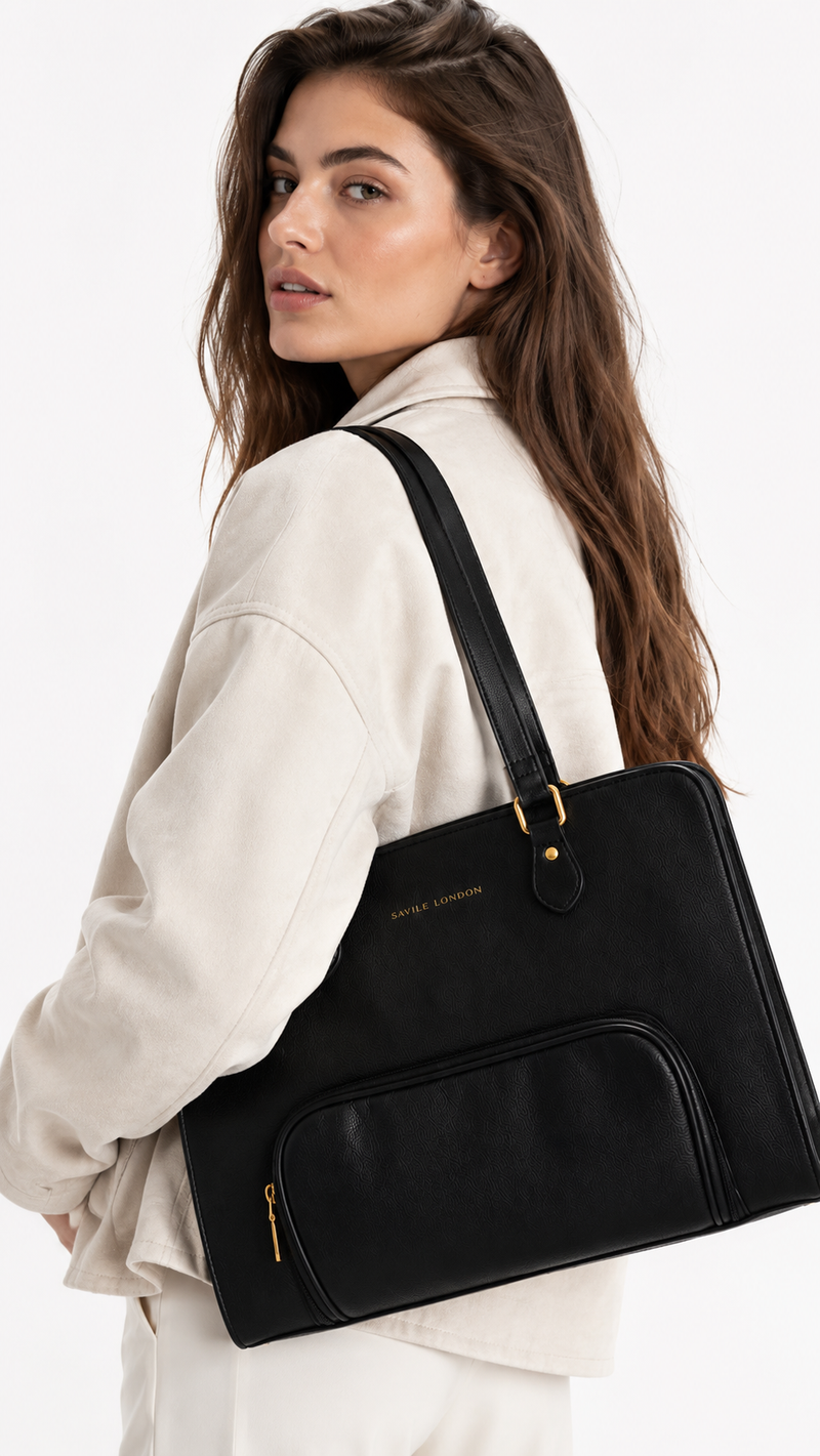 A woman wearing a cream-colored jacket and white pants carries a black leather tote bag with gold hardware and a small gold logo on the front, photographed against a clean white background.